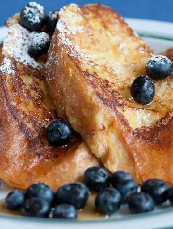 Close up view of french toast with blueberries, syrup and powdered sugar on white plate.